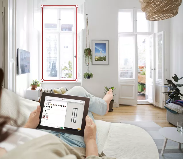 Tablette tenue par une personne allongée, consultant une application de personnalisation, dans un salon lumineux avec de grandes fenêtres ouvertes sur l'extérieur.