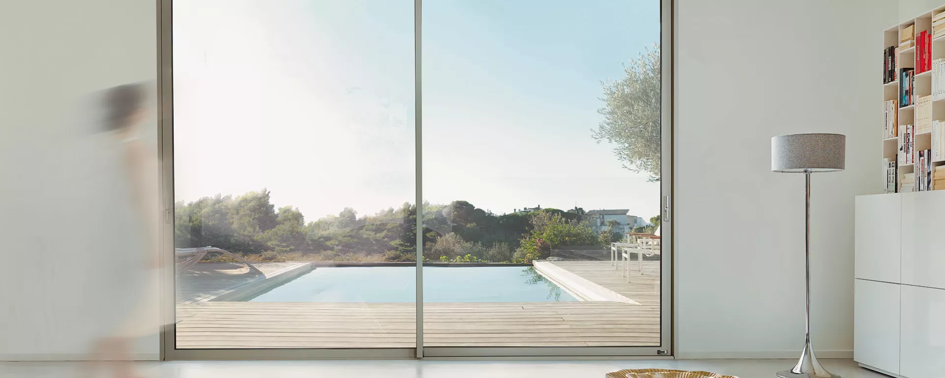 Piscine, entourée de verdure, visible à travers une immense baie vitrée dans une salle moderne avec bibliothèque et lampe. Une silhouette floue marche dans la pièce.