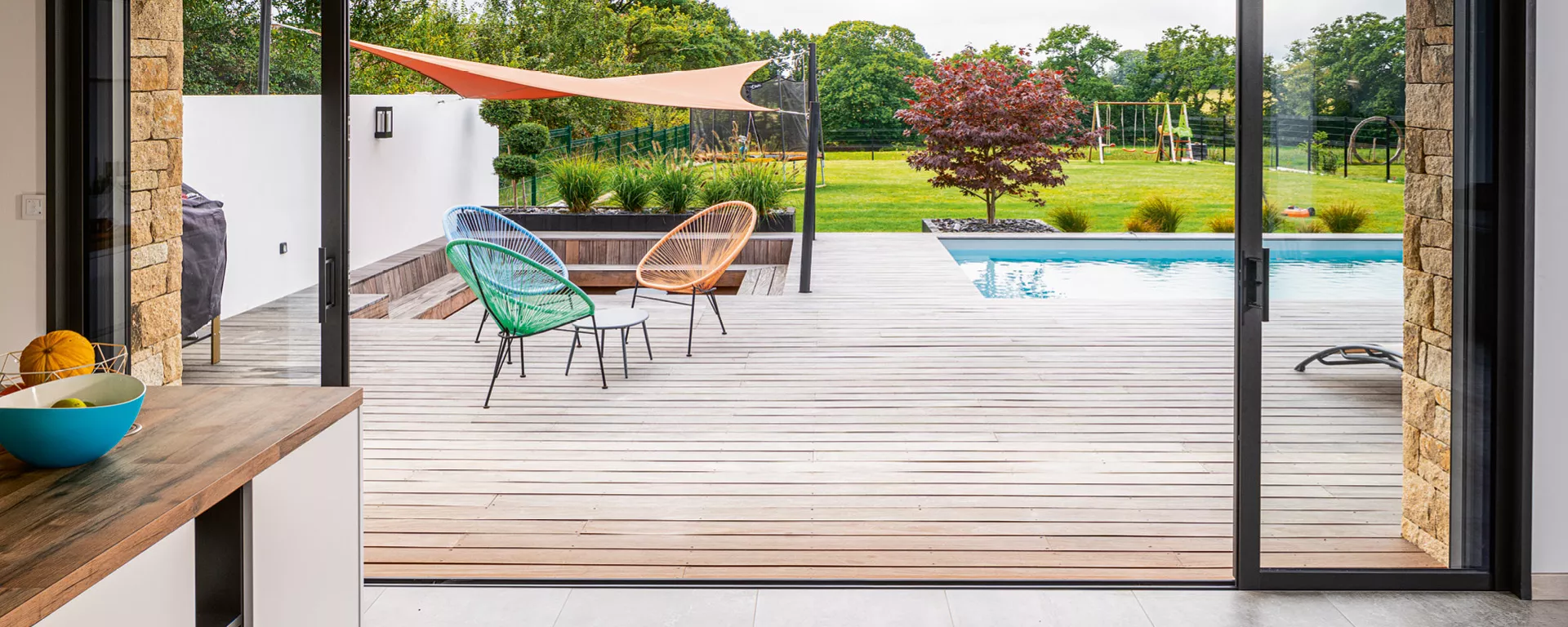 Deux chaises colorées et une table basse sur une terrasse bois, face à une piscine et un jardin verdoyant, avec un store orange.