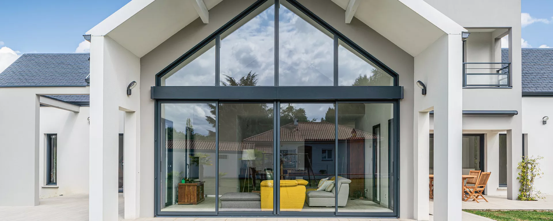 Maison moderne aux façades blanches, de larges baies vitrées donnant sur un jardin, intérieurs lumineux avec mobilier minimaliste, ciel bleu en toile de fond.