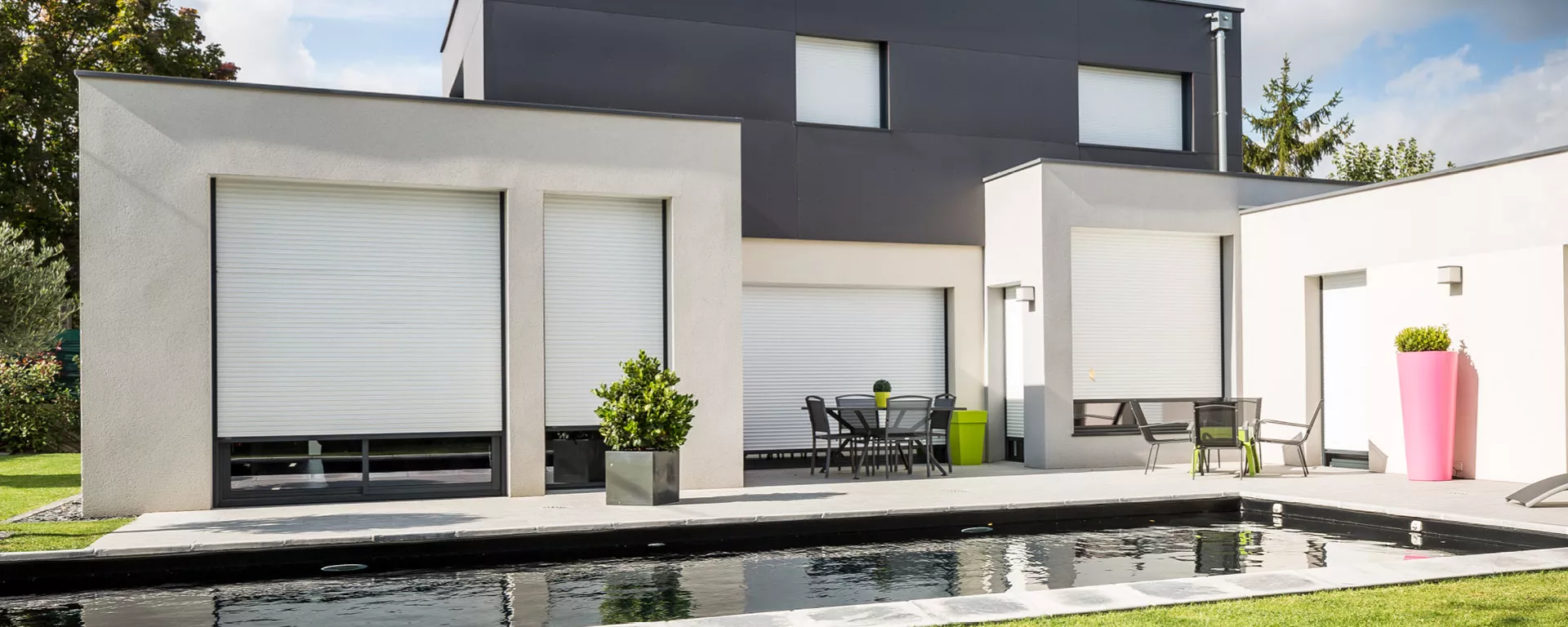 Maison moderne avec volets roulants fermés, entourant une terrasse équipée de tables et chaises, près d'une piscine rectangulaire et de pots de fleurs colorés, en extérieur.