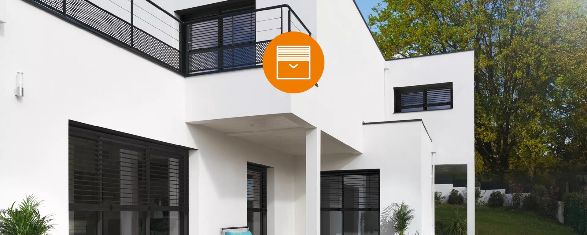 Maison moderne blanche, de larges baies vitrées, un balcon à garde-corps noir, entourée de verdure, sous un ciel dégagé.