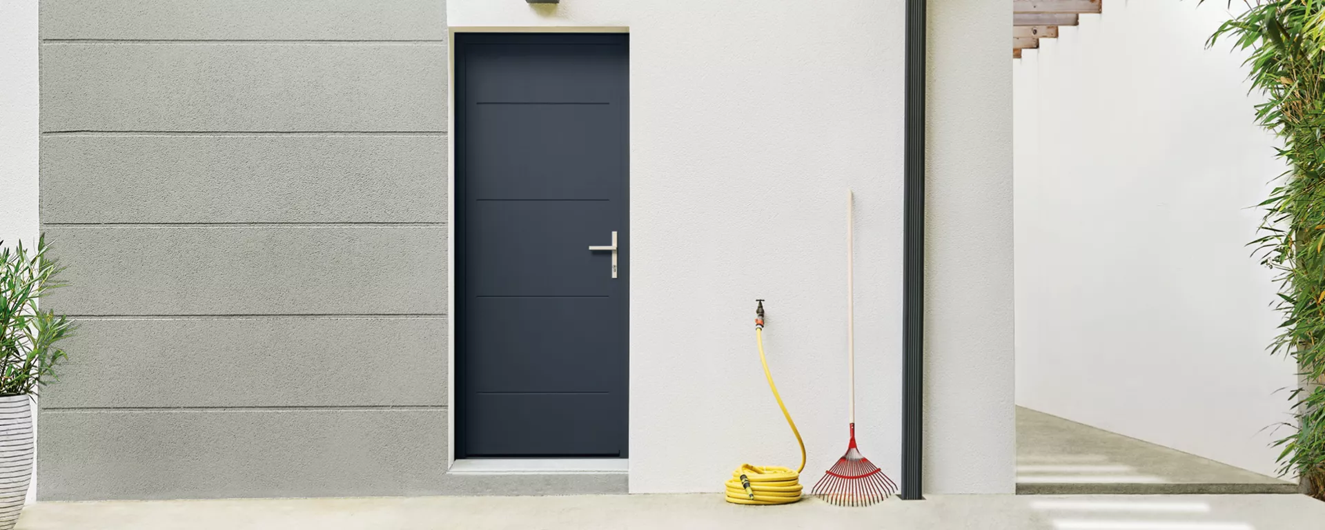 Une porte noire se trouve dans un mur blanc avec un tuyau jaune enroulé, un râteau rouge et une plante en pot à proximité.