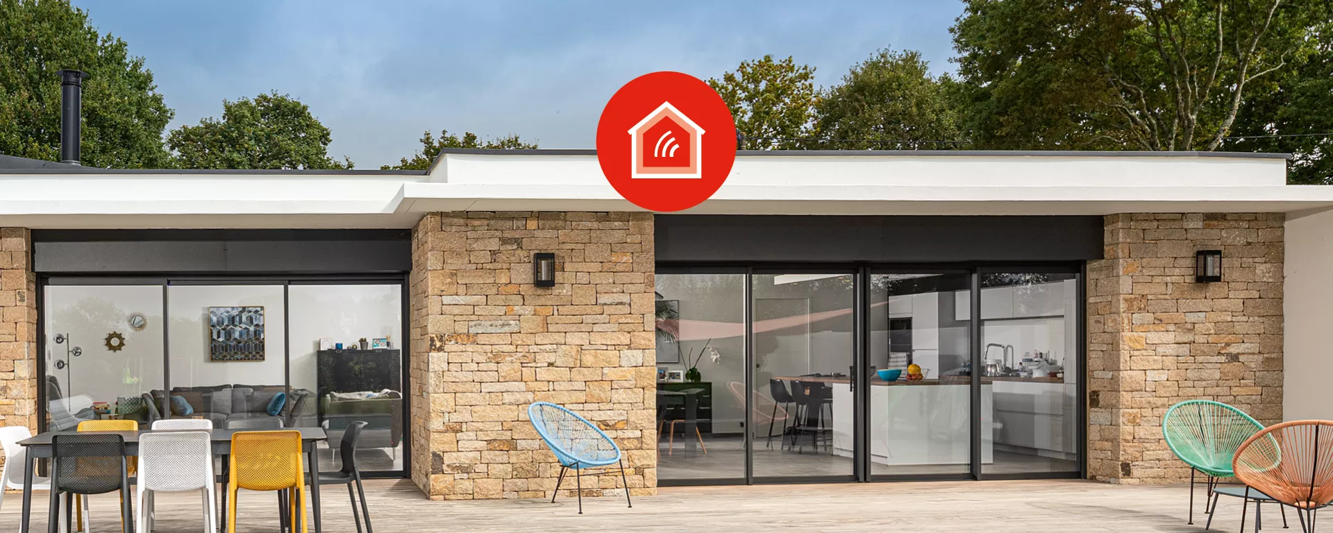 Maison moderne, fenêtres coulissantes, terrasse en bois avec chaises colorées, entourée d'arbres. Le logo d'une maison rouge avec un signal est et affiché au-dessus.