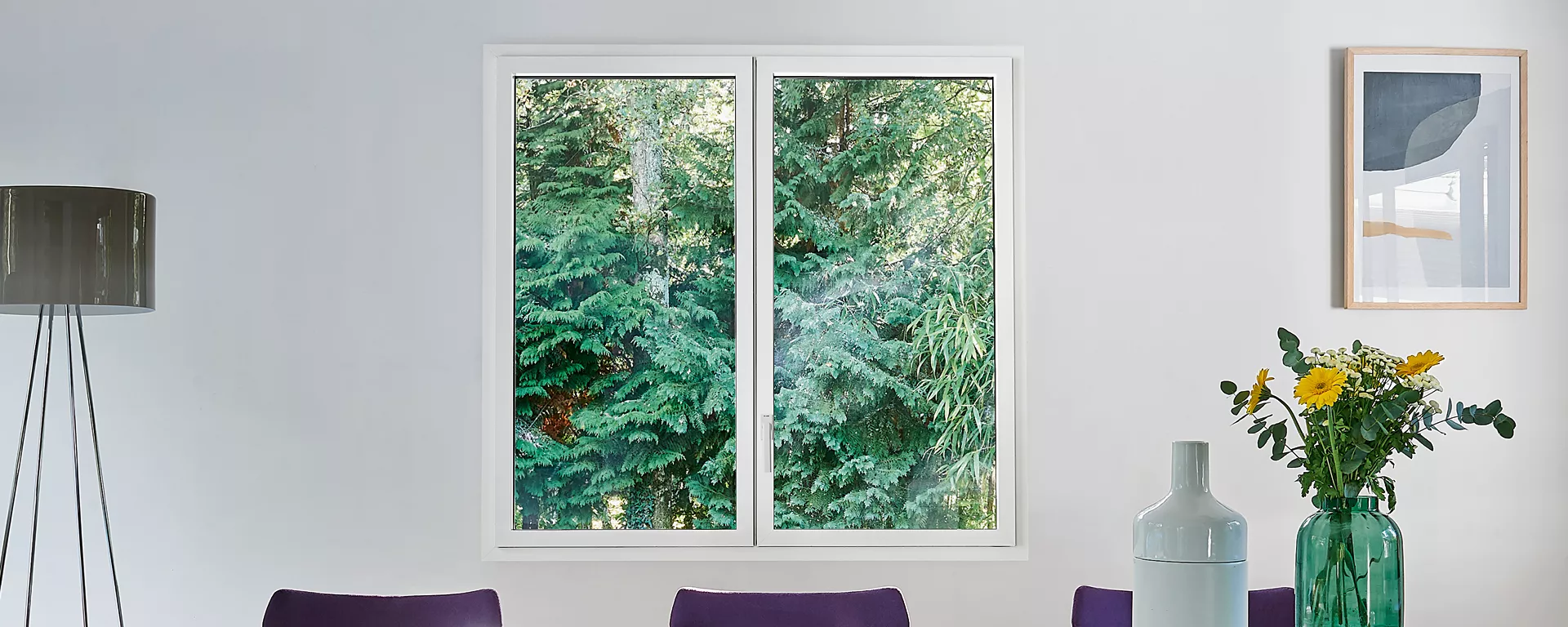 Fenêtre blanche expose des arbres verts, entourée d'un lampadaire, d'une peinture et d'un vase avec fleurs jaunes dans une pièce sobre.