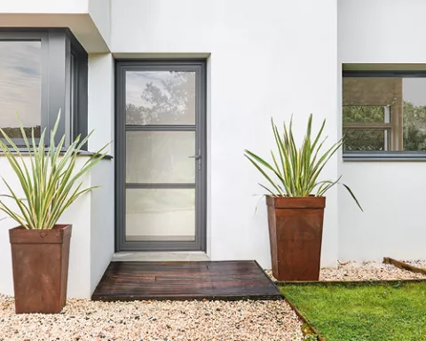 Porte en verre encadrée de gris, entourée de murs blancs, avec une plante verte en pot à droite et un chemin en graviers et bois au sol.