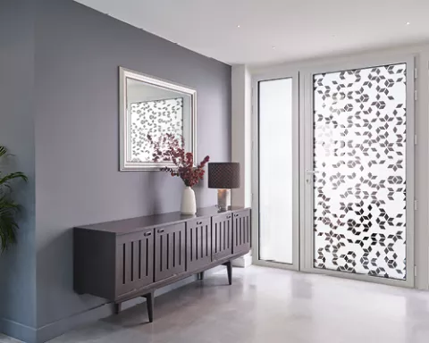 Buffet en bois avec vase et lampe, devant une fenêtre décorée de motifs floraux. Miroir encadré au-dessus, murs gris et sol clair. Atmosphère moderne et lumineuse.