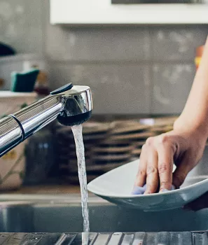 Personne lavant une assiette dans un évier avec de l'eau courante, entourée de produits vaisselle dans une cuisine moderne.