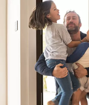 Un père porte ses deux enfants avec joie dans une entrée lumineuse, créant une scène de retrouvailles chaleureuses à la maison.