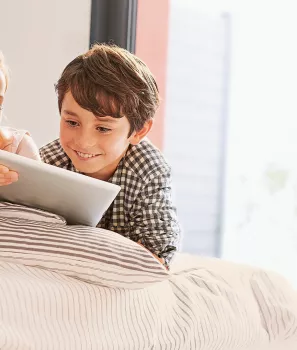 Deux enfants regardent une tablette numérique ensemble, souriant et allongés sur un lit à rayures dans une pièce lumineuse.