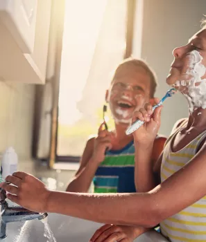 Deux enfants souriants jouent à se raser, debout devant un lavabo, avec de la mousse à raser sur leurs visages.