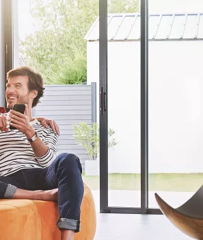 Un homme et une femme sourient et regardent un smartphone, assis sur un canapé orange dans un salon lumineux avec grande baie vitrée.