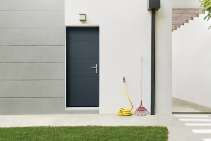 Une porte noire se trouve dans un mur blanc avec un tuyau jaune enroulé, un râteau rouge et une plante en pot à proximité.