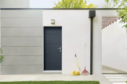 Une porte noire est encadrée par des murs blancs, avec un tuyau d'arrosage jaune et un balai posés à côté, dans un contexte extérieur.