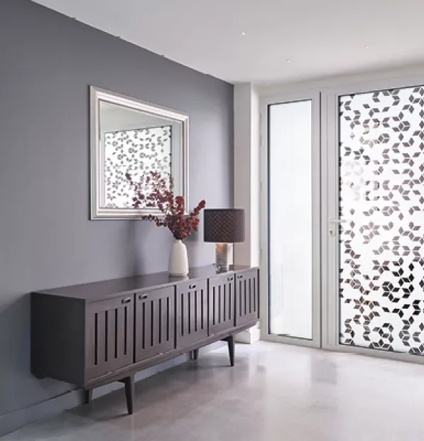 Buffet en bois avec vase et lampe, devant une fenêtre décorée de motifs floraux. Miroir encadré au-dessus, murs gris et sol clair. Atmosphère moderne et lumineuse.