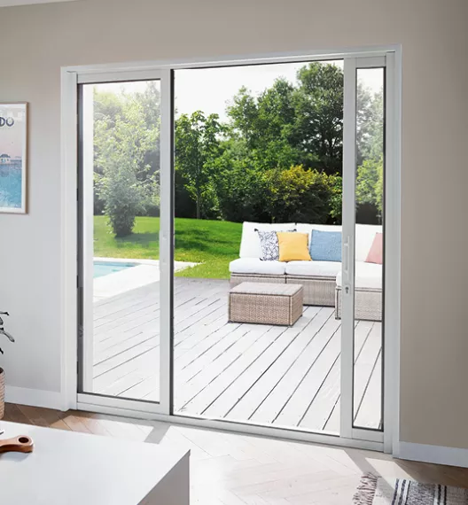 Une grande porte-fenêtre coulissante s'ouvre sur une terrasse en bois, où un canapé et une table basse sont placés, donnant sur un jardin verdoyant.